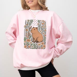 Puede incluir: Una sudadera rosa claro con una ilustraci&oacute;n de carpincho marr&oacute;n rodeada de flores y estrellas. El carpincho est&aacute; en un dise&ntilde;o floral rectangular con hojas verdes y flores naranjas y amarillas. La sudadera es de estilo casual.