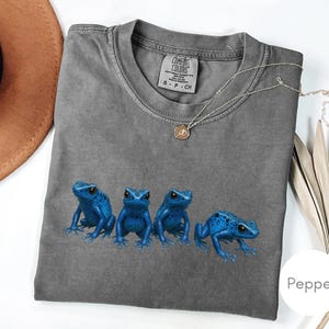 May include: A gray Comfort Colors t-shirt featuring four vibrant blue frog illustrations. The shirt has a crew neck and short sleeves. A gold necklace with a pendant is draped across the shirt.