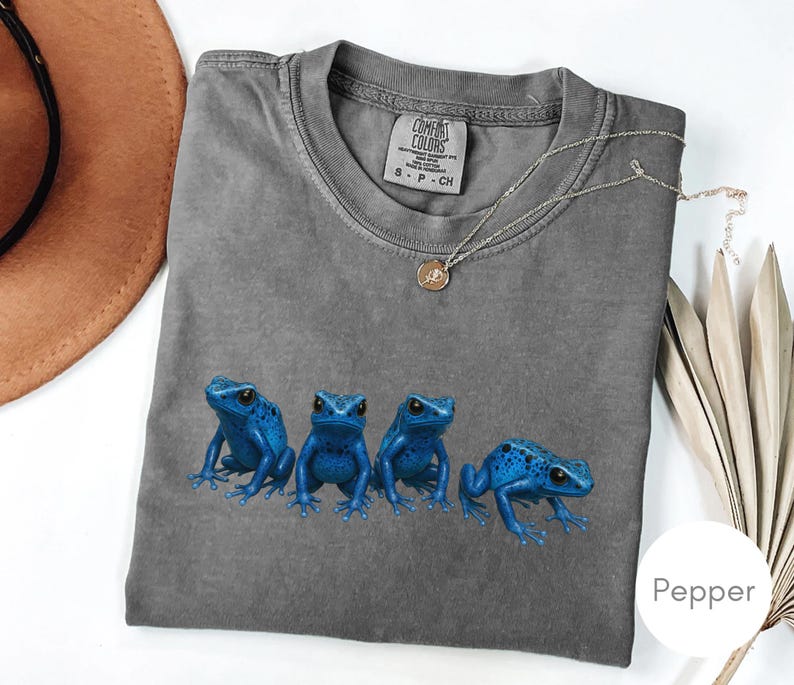 May include: A gray Comfort Colors t-shirt featuring four vibrant blue frog illustrations. The shirt has a crew neck and short sleeves. A gold necklace with a pendant is draped across the shirt.