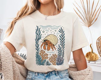 May include: A cream-coloured t-shirt with a detailed illustration of a hermit crab in a shell, surrounded by seaweed and coral. The design uses orange, blue, and beige, creating a coastal theme. The shirt is made of soft material.
