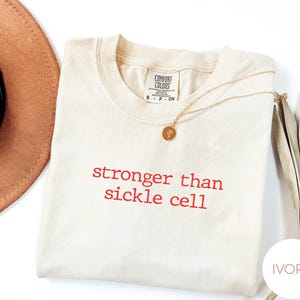 May include: Ivory-colored t-shirt with the text "stronger than sickle cell" in red. A gold necklace with a pendant rests on the shirt. A tan hat and dried plant stems are in the background.