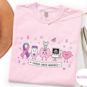 May include: A light pink t-shirt featuring a graphic print of cartoon characters including a ribbon, a pill bottle, a thyroid gland, an ultrasound machine, and a heart. Below the characters is a banner with the text THYROID CANCER AWARENESS.