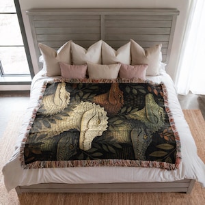 May include: A woven throw blanket featuring a repeating pattern of dinosaur heads in shades of cream, brown, and green against a dark background. The blanket has a fringed edge and is displayed on a bed with pillows.