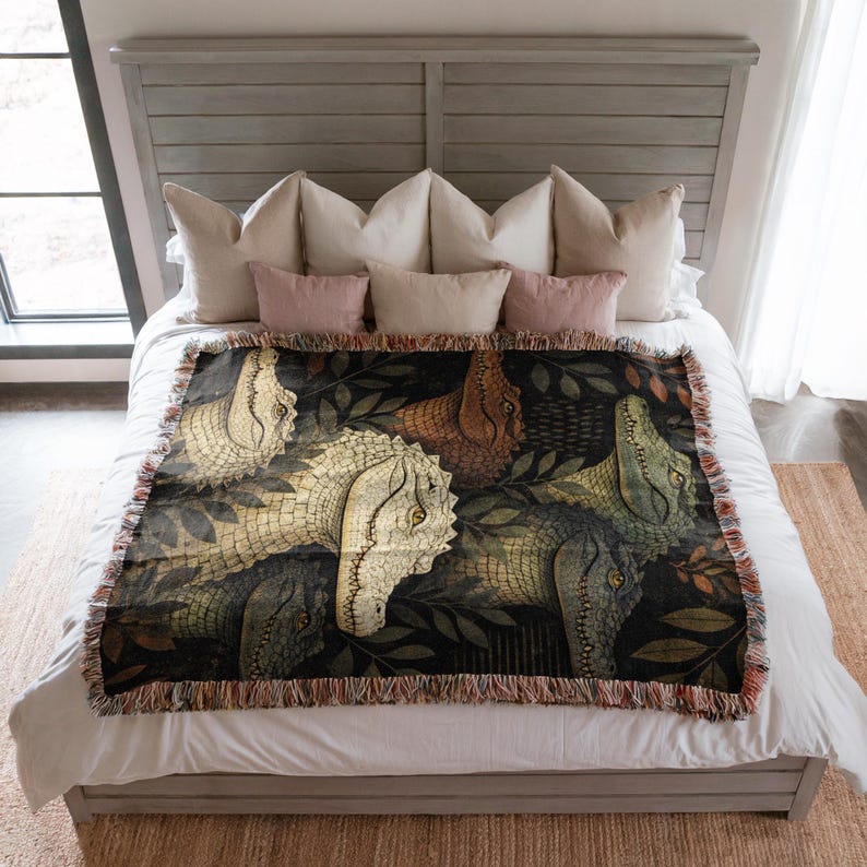 May include: A woven throw blanket featuring a repeating pattern of dinosaur heads in shades of cream, brown, and green against a dark background. The blanket has a fringed edge and is displayed on a bed with pillows.