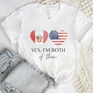 Può includere: T-shirt bianco con un design a forma di cuore raffigurante la bandiera peruviana e la bandiera americana. Il testo "YES, I'M BOTH of them" è stampato sotto. La maglietta è abbinata a jeans blu.