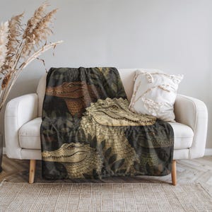 May include: A throw blanket featuring a detailed illustration of several alligators in a jungle setting. The alligators are depicted in shades of brown and beige, with a dark green background of foliage. The blanket is draped over a white sofa.