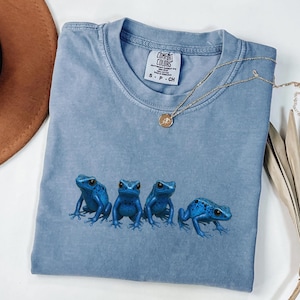 May include: A light blue Comfort Colors t-shirt featuring four blue frog illustrations. The shirt has a crew neck and short sleeves. A gold necklace with a pendant rests on the shirt. The shirt is folded.