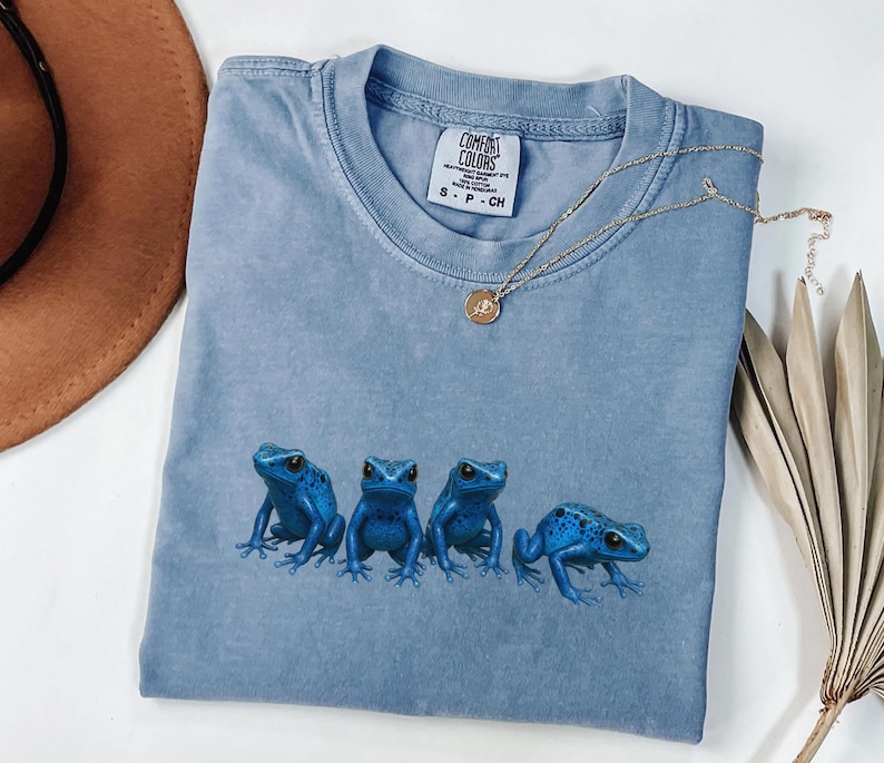 May include: A light blue Comfort Colors t-shirt featuring four blue frog illustrations. The shirt has a crew neck and short sleeves. A gold necklace with a pendant rests on the shirt. The shirt is folded.