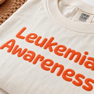 May include: An off-white crew neck t-shirt featuring the text "Leukemia Awareness" printed in bold, raised orange lettering across the chest. The garment includes a Comfort Colors brand label inside the collar area near the neckline.