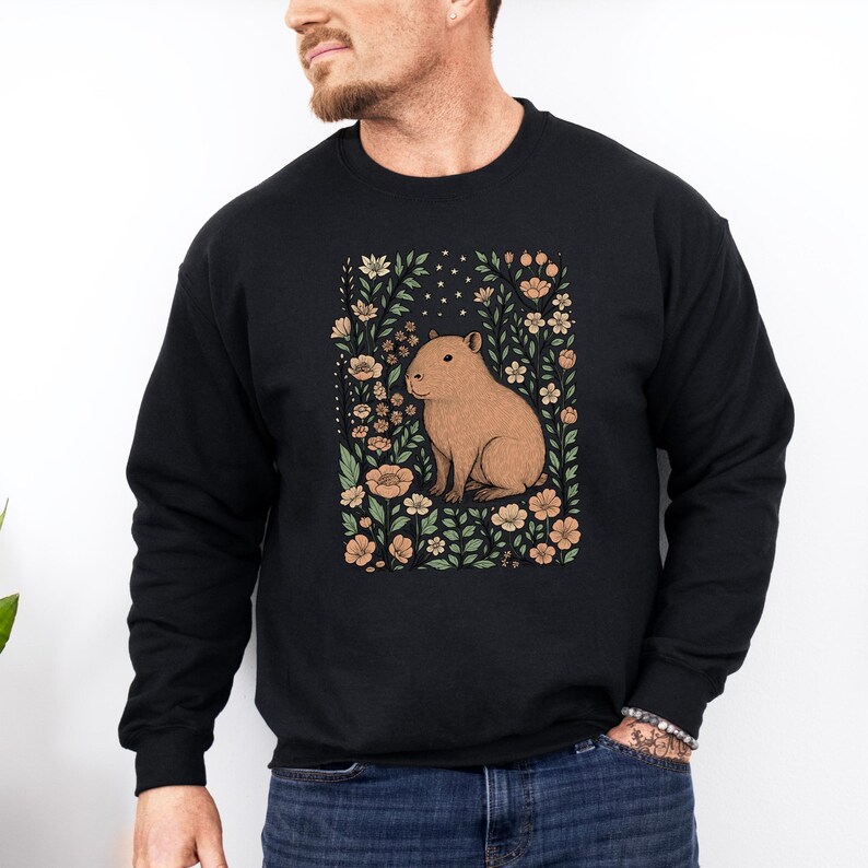 Puede incluir: Sudadera negra con una ilustraci&oacute;n de carpincho marr&oacute;n rodeada de flores y follaje en tonos melocot&oacute;n, verde y crema. El dise&ntilde;o incluye peque&ntilde;as estrellas. La sudadera es de cuello redondo.