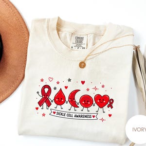 May include: An ivory t-shirt featuring a graphic of five red cartoon characters holding hands, including a ribbon, a blood drop, a crescent, a circle, and a heart. Below the characters is a banner with the text SICKLE CELL AWARENESS.