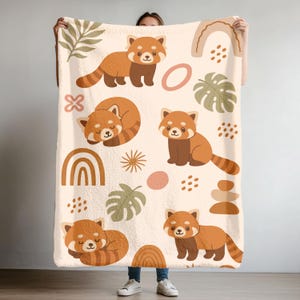 May include: A cream-colored blanket featuring a repeating pattern of cartoon red pandas in various poses. The design includes brown rainbows, leaves, and geometric shapes in shades of brown and pink. The blanket is soft and cozy.