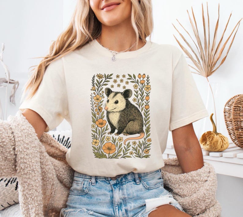 May include: Cream-colored t-shirt featuring a detailed illustration of an opossum within a floral frame. The opossum is depicted in shades of gray, brown, and cream, surrounded by orange and green floral elements. The shirt is a casual wear item.
