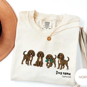 May include: An ivory-colored short-sleeve t-shirt featuring a graphic of four brown brindle dogs. One dog holds a teal toy. Below the dogs, the text reads "Dog name (optional)". The shirt has a Comfort Colors brand label at the inner neckline.
