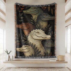 May include: Woven throw blanket featuring a detailed illustration of several alligators in various colors, including green, brown, and cream. The alligators are surrounded by dark green leaves against a black background. The blanket has a fringed edge.