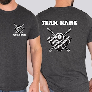 Könnte beinhalten: Dunkelgraues T-Shirt mit weißer Billardkugel-Grafik auf Vorder- und Rückseite. Die Vorderseite zeigt gekreuzte Queues und einen Satz Kugeln mit dem Text "PLAYER NAME". Die Rückseite zeigt "TEAM NAME" über einem ähnlichen Billardkugel-Design.