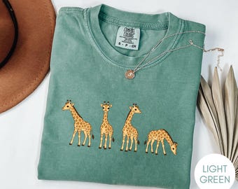 May include: A light green short-sleeve t-shirt featuring a graphic of four giraffes in various poses. The shirt has a crew neckline and a Comfort Colors brand label printed on the inside collar. The giraffes are brown and tan with spots.