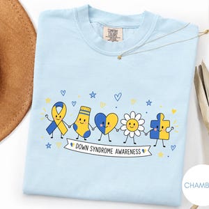 May include: A light blue t-shirt featuring a graphic of five cartoon characters holding hands: a yellow and blue awareness ribbon, a striped sock, a heart, a daisy, and a puzzle piece. Below them is a banner with the text DOWN SYNDROME AWARENESS.