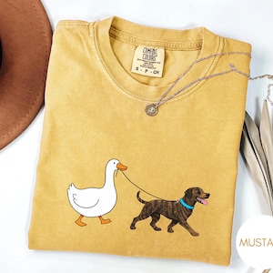 以下が含まれることがあります： マスタード色のTシャツ。アヒルが茶色の犬をリードで散歩させている漫画が描かれています。シャツにはComfort Colorsのタグとシルバーのネックレスが付いています。「MUSTARD」の文字が右下の白い円の中にあります。