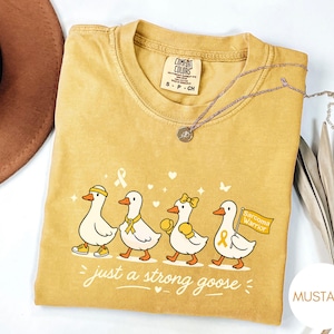 以下が含まれることがあります： マスタード色のTシャツ。4羽の漫画のガチョウが描かれており、それぞれアクセサリーを身につけている。「just a strong goose」の文字が下にあります。シルバーのネックレスがTシャツの上にかけられています。サイズS-P-CH。