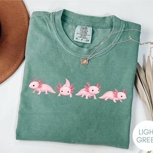 Könnte beinhalten: Ein salbeigrünes T-Shirt mit einem Grafikdruck von vier rosa Axolotln in verschiedenen Posen auf der Brust. Das T-Shirt hat einen Rundhalsausschnitt und ein Comfort Colors Markenetikett im Innenkragen, das die Größe S angibt.