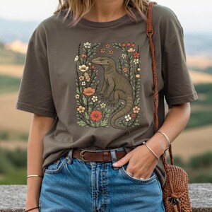May include: Olive green t-shirt featuring a detailed illustration of a lizard surrounded by flowers. The artwork includes a variety of blooms in shades of orange, white, and yellow, set against a backdrop of green foliage. The shirt has short sleeves and a crew neck.