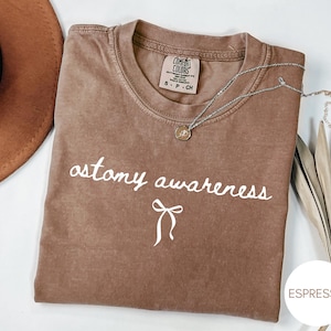 May include: Espresso-colored t-shirt with the words "ostomy awareness" in white script, along with a white bow graphic. A silver necklace with a small pendant rests on the shirt. The shirt is folded, and a brown hat is visible in the background.