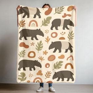 May include: A cream-colored blanket featuring a repeating pattern of brown tapirs, leaves, rainbows, and abstract shapes. The blanket has a soft, plush texture, perfect for snuggling. The design is a whimsical, nature-inspired theme.