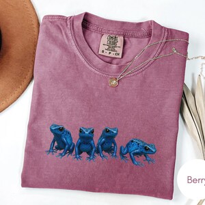 May include: Berry-colored t-shirt featuring a crew neck and short sleeves. The shirt has a graphic of four blue frogs. A gold necklace with a pendant is draped across the shirt. The shirt is labeled "Comfort Colors".