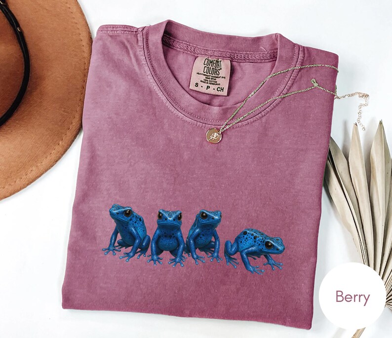May include: Berry-colored t-shirt featuring a crew neck and short sleeves. The shirt has a graphic of four blue frogs. A gold necklace with a pendant is draped across the shirt. The shirt is labeled "Comfort Colors".