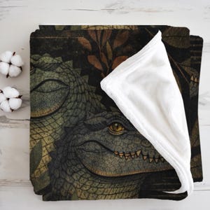 May include: A folded blanket featuring a detailed illustration of two alligators with yellow eyes and open mouths. The blanket has a dark background with green and brown foliage. The underside of the blanket is white.