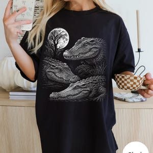 May include: A black t-shirt featuring a detailed white graphic print of three alligators in a swamp setting under a full moon. The design showcases the textured scales of the reptiles and the silhouette of a tree against the circular moon.