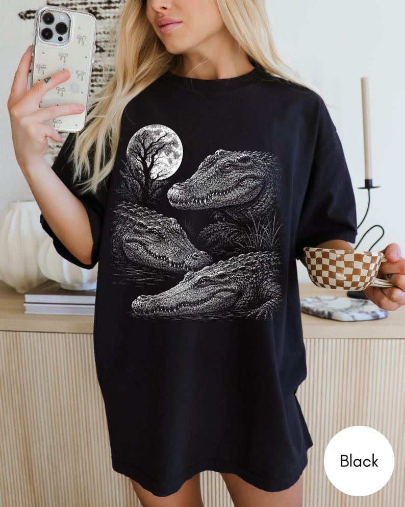 May include: A black t-shirt featuring a detailed white graphic print of three alligators in a swamp setting under a full moon. The design showcases the textured scales of the reptiles and the silhouette of a tree against the circular moon.