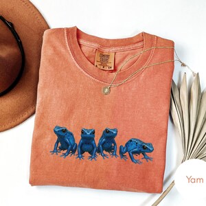 May include: A coral-colored t-shirt featuring four blue frog illustrations. The shirt has a crew neck and short sleeves. A gold necklace with a pendant is draped across the shirt. A brown hat and a dried plant are in the background.