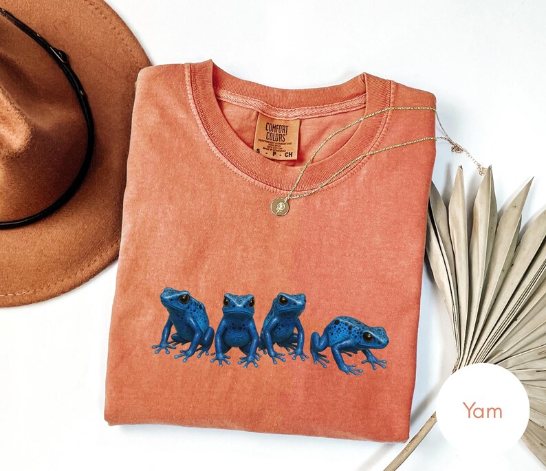 May include: A coral-colored t-shirt featuring four blue frog illustrations. The shirt has a crew neck and short sleeves. A gold necklace with a pendant is draped across the shirt. A brown hat and a dried plant are in the background.