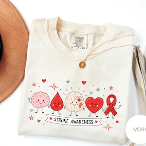 Könnte beinhalten: Ein elfenbeinfarbenes T-Shirt mit einer Grafik von fünf Cartoon-Figuren, die sich an den Händen halten: ein rosa Gehirn, ein roter Blutstropfen, ein beiges Gehirn mit rotem Kreis, ein rotes Herz und eine rote Schleife mit dem Text STROKE AWARENESS.