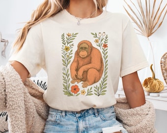 Puede incluir: Camiseta color crema con una ilustración de un orangután enmarcada por flores y hojas. El orangután está representado en tonos naranja y marrón, con follaje verde y flores coloridas. La camiseta tiene mangas cortas y un corte relajado.