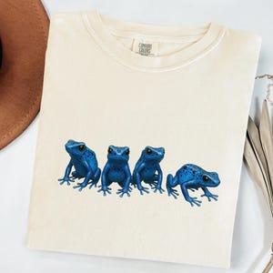 May include: Cream-colored t-shirt featuring a graphic of four vibrant blue frogs. The frogs are detailed with black eyes and spots, arranged in a row across the shirt. The shirt has a crew neck and short sleeves.