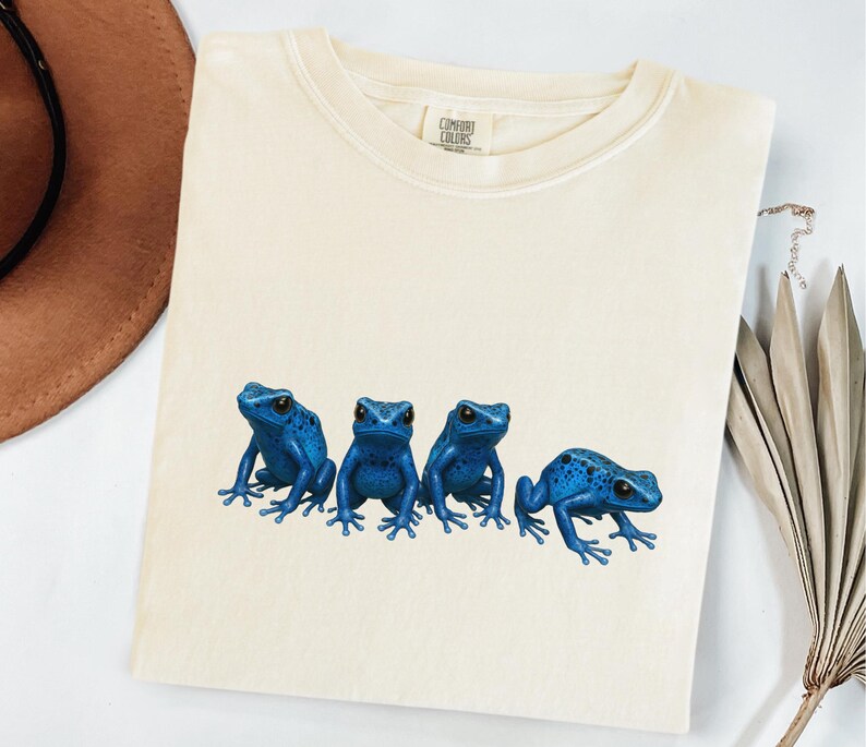 May include: Cream-colored t-shirt featuring a graphic of four vibrant blue frogs. The frogs are detailed with black eyes and spots, arranged in a row across the shirt. The shirt has a crew neck and short sleeves.