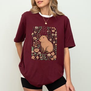 Puede incluir: Camiseta burdeos con una ilustraci&oacute;n de carpincho rodeado de flores. El carpincho es de color marr&oacute;n claro y las flores son melocot&oacute;n y blancas. La camiseta tiene cuello blanco y corte holgado.