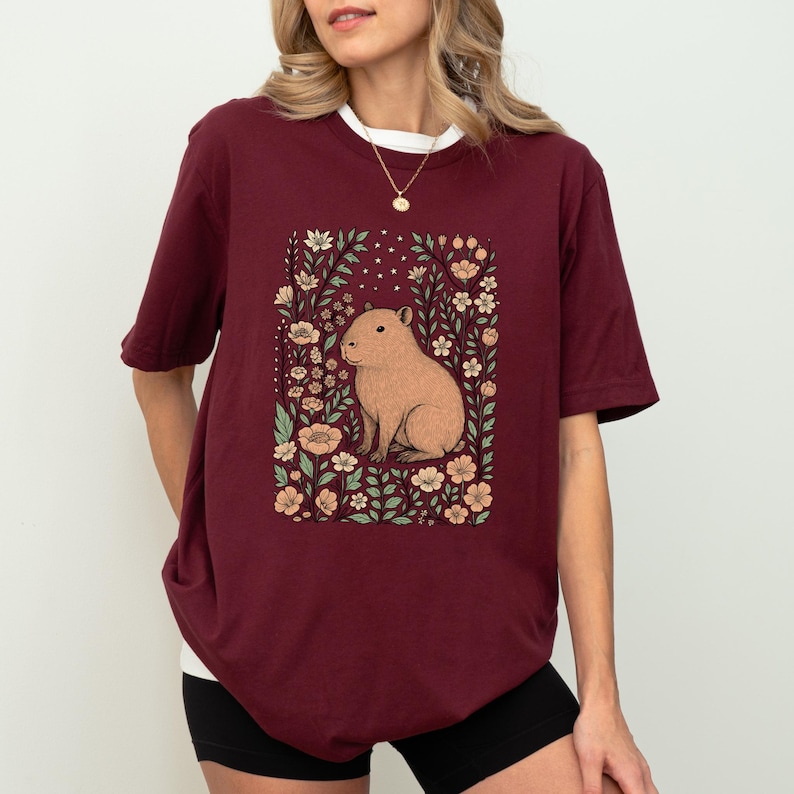 Puede incluir: Camiseta burdeos con una ilustraci&oacute;n de carpincho rodeado de flores. El carpincho es de color marr&oacute;n claro y las flores son melocot&oacute;n y blancas. La camiseta tiene cuello blanco y corte holgado.