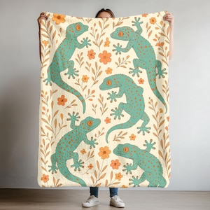 May include: A cream-colored blanket featuring a repeating pattern of teal geckos with orange spots and eyes, interspersed with orange flowers and brown leaves. The blanket is held up by a person.