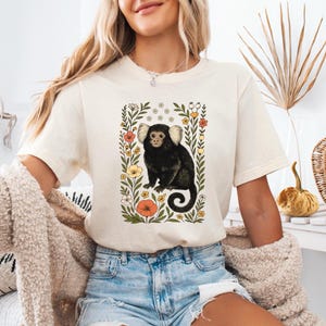 Może przedstawiać: Kremowy t-shirt z ilustracją czarnej małpy otoczonej kolorowymi kwiatami i zielonymi liśćmi. Małpa ma białe pęczki uszu i zakręcony ogon. Projekt jest wyśrodkowany, tworząc botaniczną i zwierzęcą estetykę.