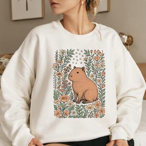 Puede incluir: Sudadera crema con una ilustraci&oacute;n de carpincho rodeado de flores y estrellas. El carpincho es marr&oacute;n claro, las flores son melocot&oacute;n y crema, y el dise&ntilde;o est&aacute; enmarcado por hojas verdes. Una prenda de vestir c&oacute;moda y con estilo.