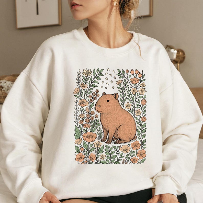 Puede incluir: Sudadera crema con una ilustraci&oacute;n de carpincho rodeado de flores y estrellas. El carpincho es marr&oacute;n claro, las flores son melocot&oacute;n y crema, y el dise&ntilde;o est&aacute; enmarcado por hojas verdes. Una prenda de vestir c&oacute;moda y con estilo.