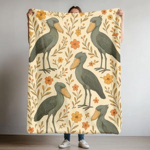 May include: A beige blanket featuring a repeating pattern of Shoebill storks, orange flowers, and brown leaves. The storks are gray with large, tan beaks. The flowers and leaves are in a retro style.
