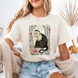 May include: Cream-colored t-shirt featuring a detailed illustration of a capuchin monkey within a floral frame. The monkey is depicted with a white face and dark brown body, surrounded by colorful flowers and green foliage. The shirt is a casual, everyday wear item.