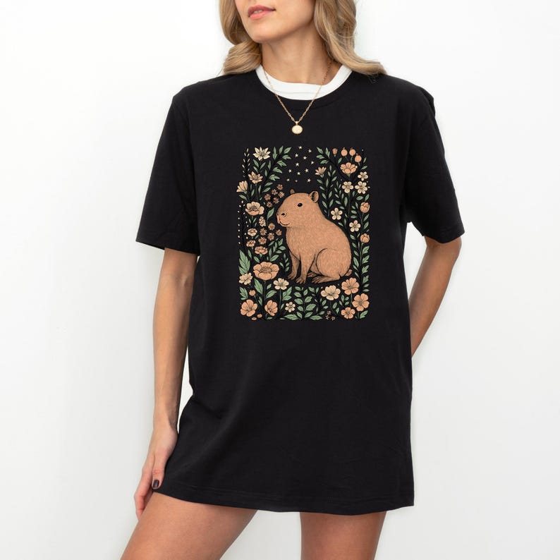 Puede incluir: Camiseta negra con una ilustraci&oacute;n de carpincho rodeado de flores. El carpincho es marr&oacute;n y las flores son melocot&oacute;n y verdes. La camiseta tiene cuello blanco y la lleva una persona.