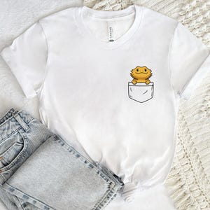 Peut inclure: T-shirt blanc avec un motif de poche représentant un dessin animé de dragon barbu. Le lézard est jaune et marron, sortant d'une poche blanche. Le t-shirt est présenté avec un jean en denim bleu clair.