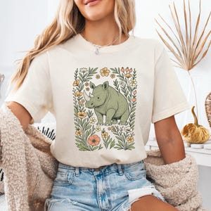 May include: Cream-colored t-shirt featuring a green rhinoceros illustration surrounded by a floral frame with yellow and peach flowers. The rhino has a white horn. The shirt has short sleeves and a relaxed fit.